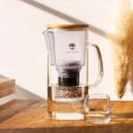 The Lotus Vita ENYA glass water filter pitcher with a bamboo lid, sitting in the sun on a wooden table next to a freshly poured glass of water.