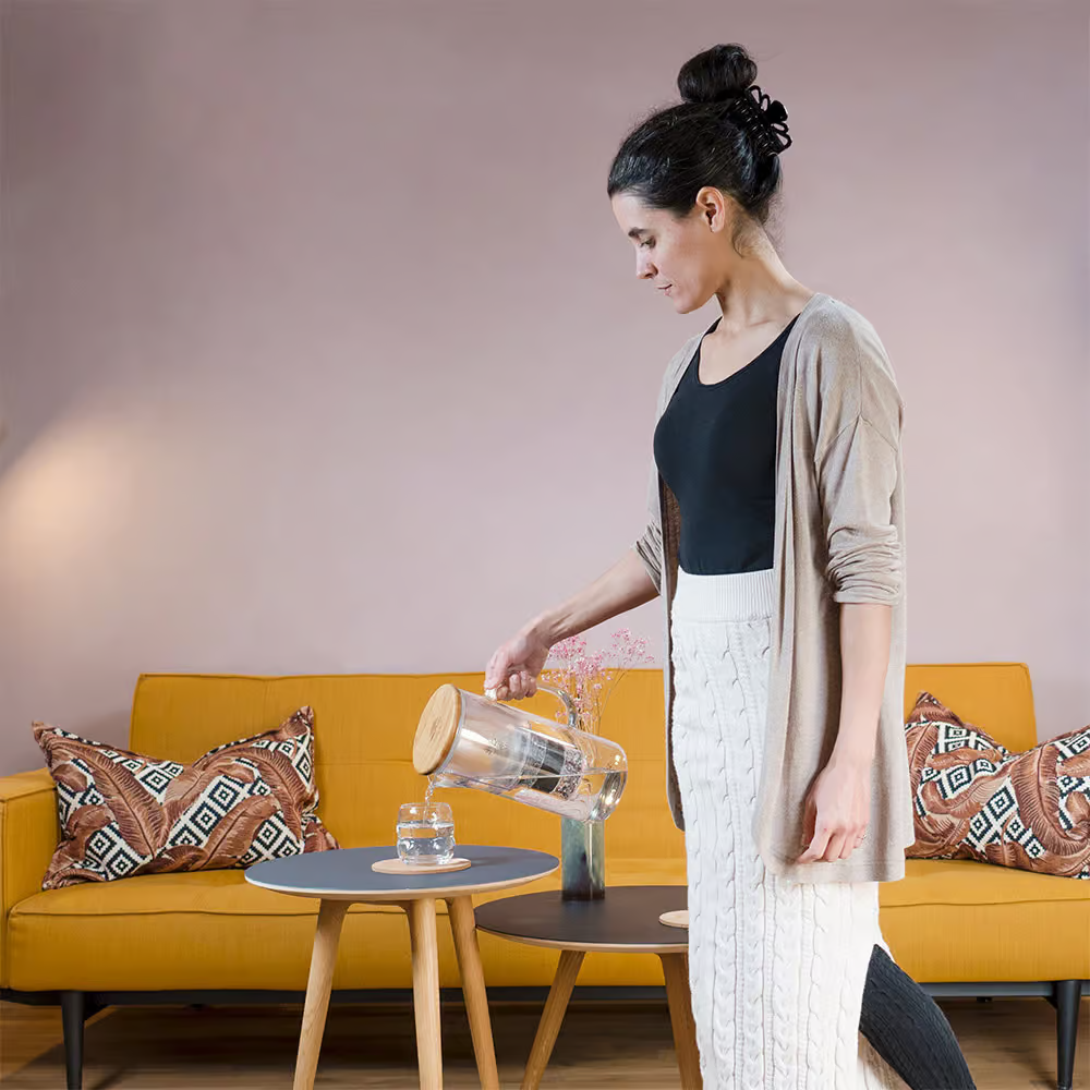 A smiling woman pours fresh, clean water from a Lotus Vita ENYA glass water filter pitcher into a drinking glass in her kitchen.