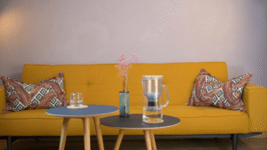 The Lotus Vita ENYA water filter pitcher on a coffee table in a modern living room, with a glass of filtered water nearby.